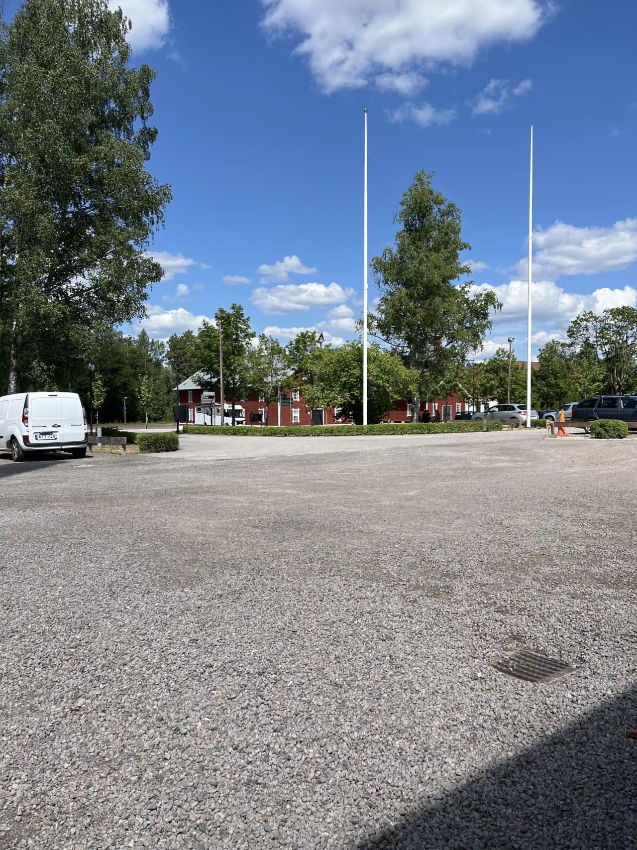 En asfalterad väg med parkeringsplats omgiven av träd, en gatlykta står vid vägkanten. Solen skapar skuggor över marken.