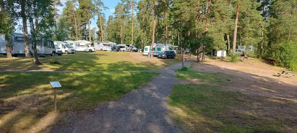 Ett campingområde med flera husbilar och husvagnar uppställda bland träden, samt en parkeringsplats för besökare.