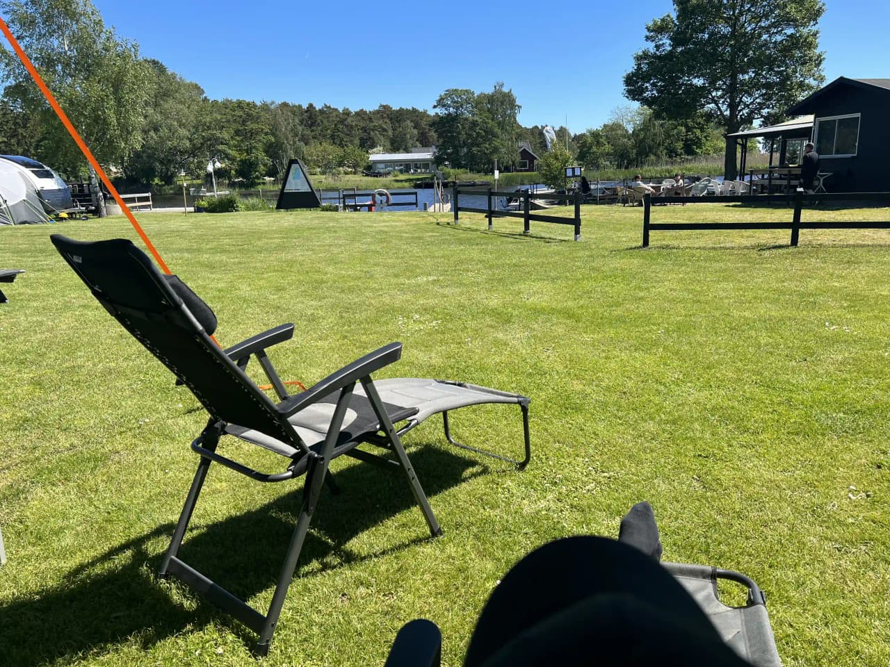 En solig dag på Martins Rökeri och Camping med utemöbler som bord och hopfällbara stolar utspridda på den gröna gräsmattan, perfekt för avkoppling. Skugga från träden ger en fridfull atmosfär.