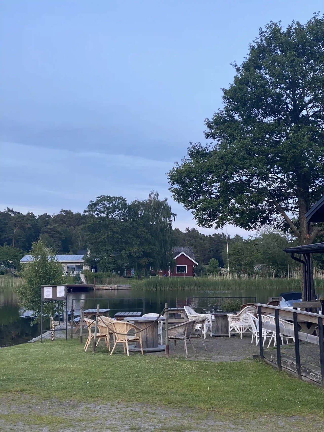 En mysig campingmiljö vid Martins Rökeri och Camping med träd, gröna växter och utomhusmöbler på en välskött gräsmatta. En stuga skymtas i bakgrunden under skuggande träd.