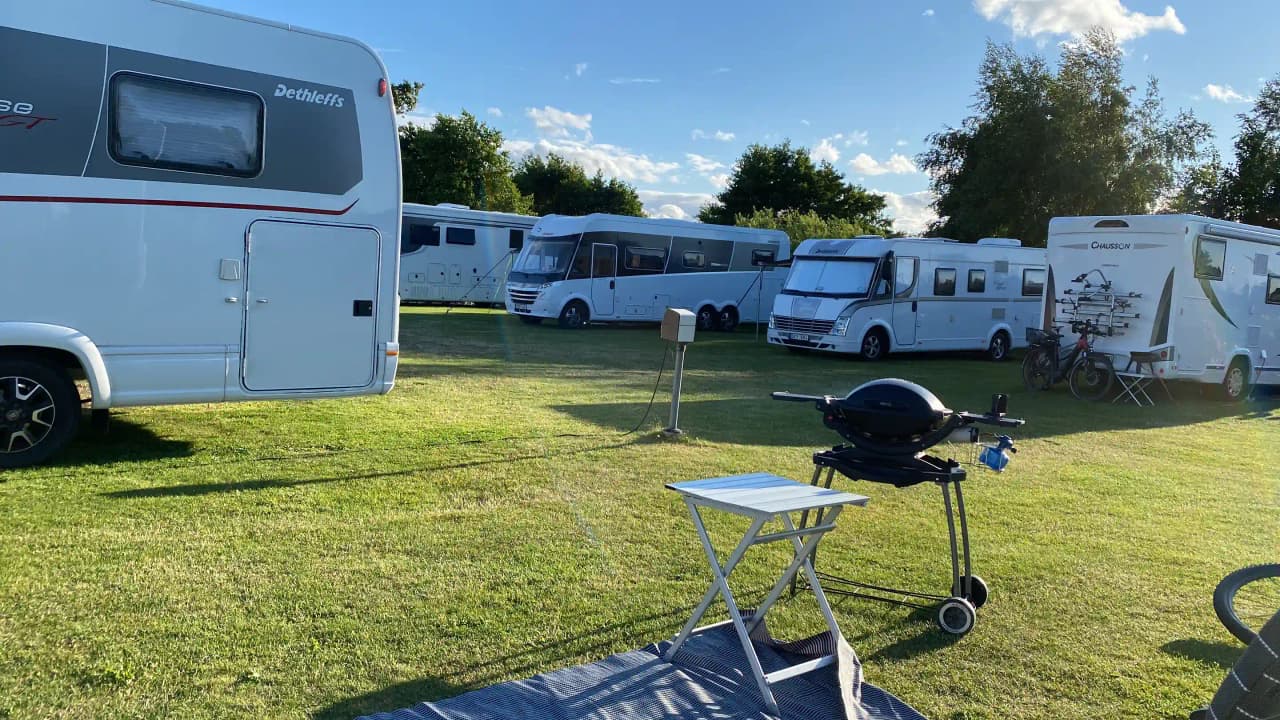 En bild av en campingplats vid Martins Rökeri och Camping, med flera husbilar och husvagnar parkerade på en gräsyta. Tält och andra campingutrustningar syns också bland träden i bakgrunden.