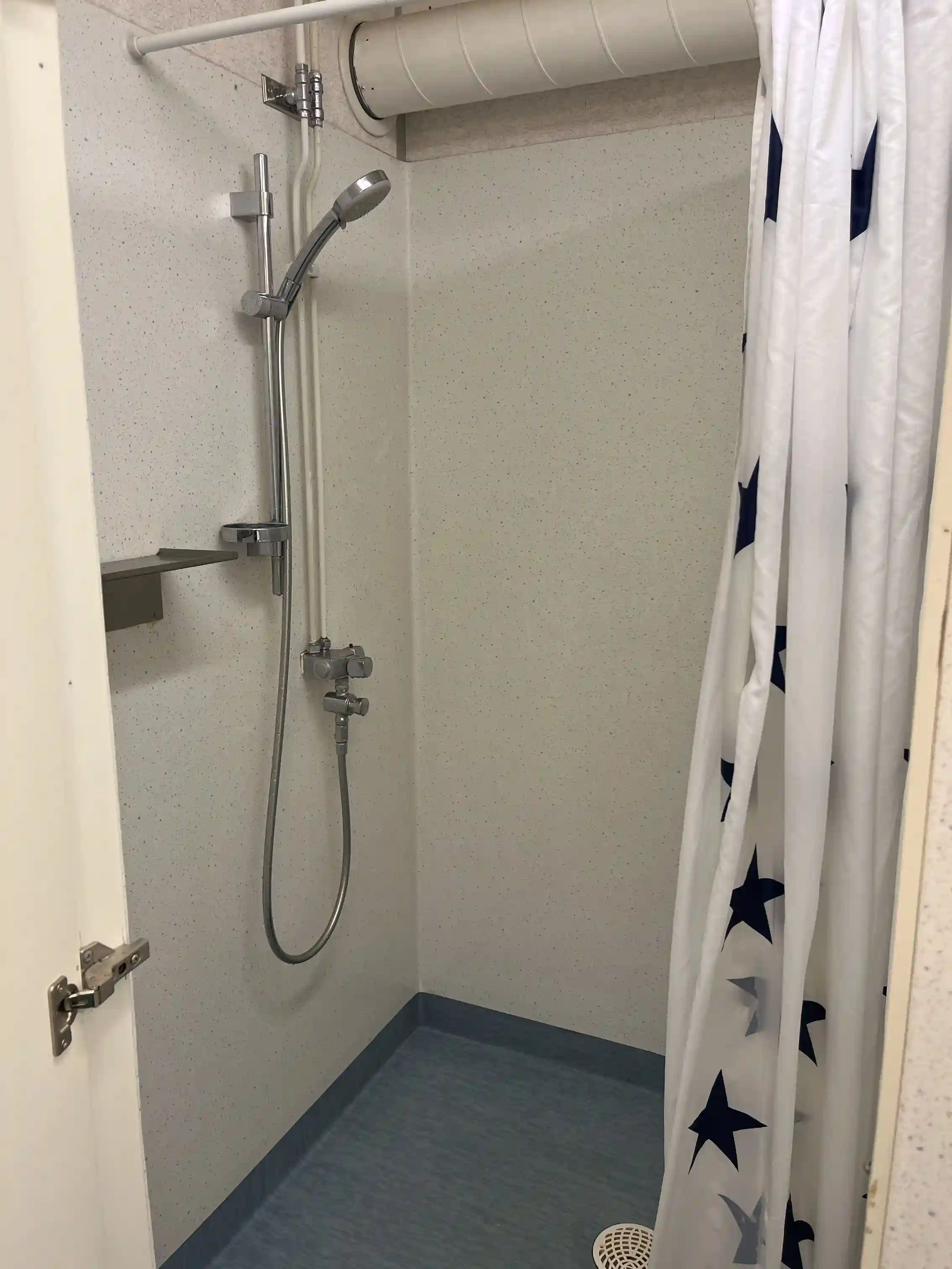 Badrum med duschområde utrustat med silverfärgat duschhuvud och duschdraperi i ljusa färger. Golvet är kaklat och rörledningar är synliga.