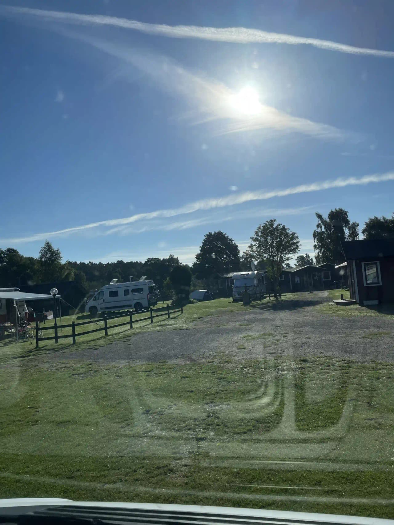 En klar himmel med några cumulusmoln ovanför en campingplats. Flera husbilar och husvagnar står parkerade på en grusplan, vilket ger en känsla av ett aktivt rekreationsområde. I bakgrunden syns öppna fält och en liten väg, vilket understryker platsens lantliga och avkopplande atmosfär.