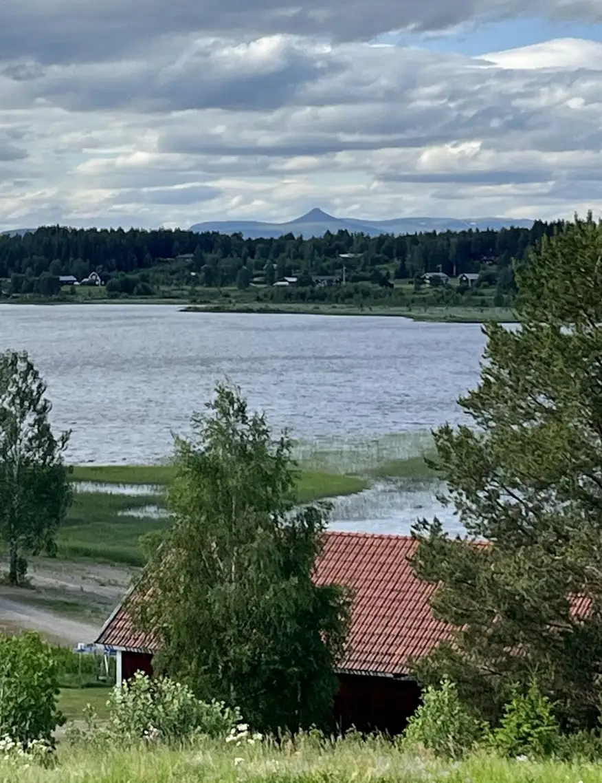 En naturskön campingplats vid en sjö med grönskande växtlighet. Moln sveper över himlen och landskapet består av vidsträckt gräsmark.
