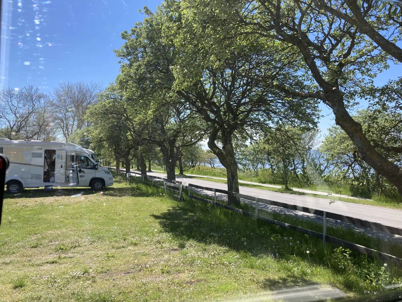 En asfalterad väg omgiven av grönska, med flera parkerade bilar av olika modeller, inklusive lyxiga fordon, som syns på vägen. Platsen är Kneippbyn Resort Visby, en populär besöksattraktion.