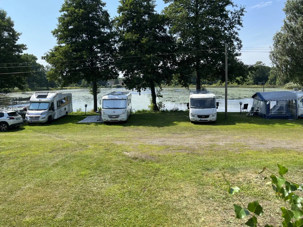 En campingplats med grön gräsmatta där flera husbilar och husvagnar är parkerade. Flera fordon står uppställda i en parkeringsliknande formation och området har en fridfull atmosfär med omgivande träd och buskar i bakgrunden.