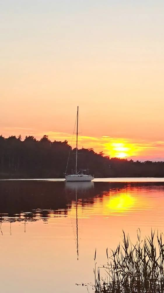 En segelbåt ligger för ankar vid vattnet i skymningen, med en vacker solnedgång vid horisonten.