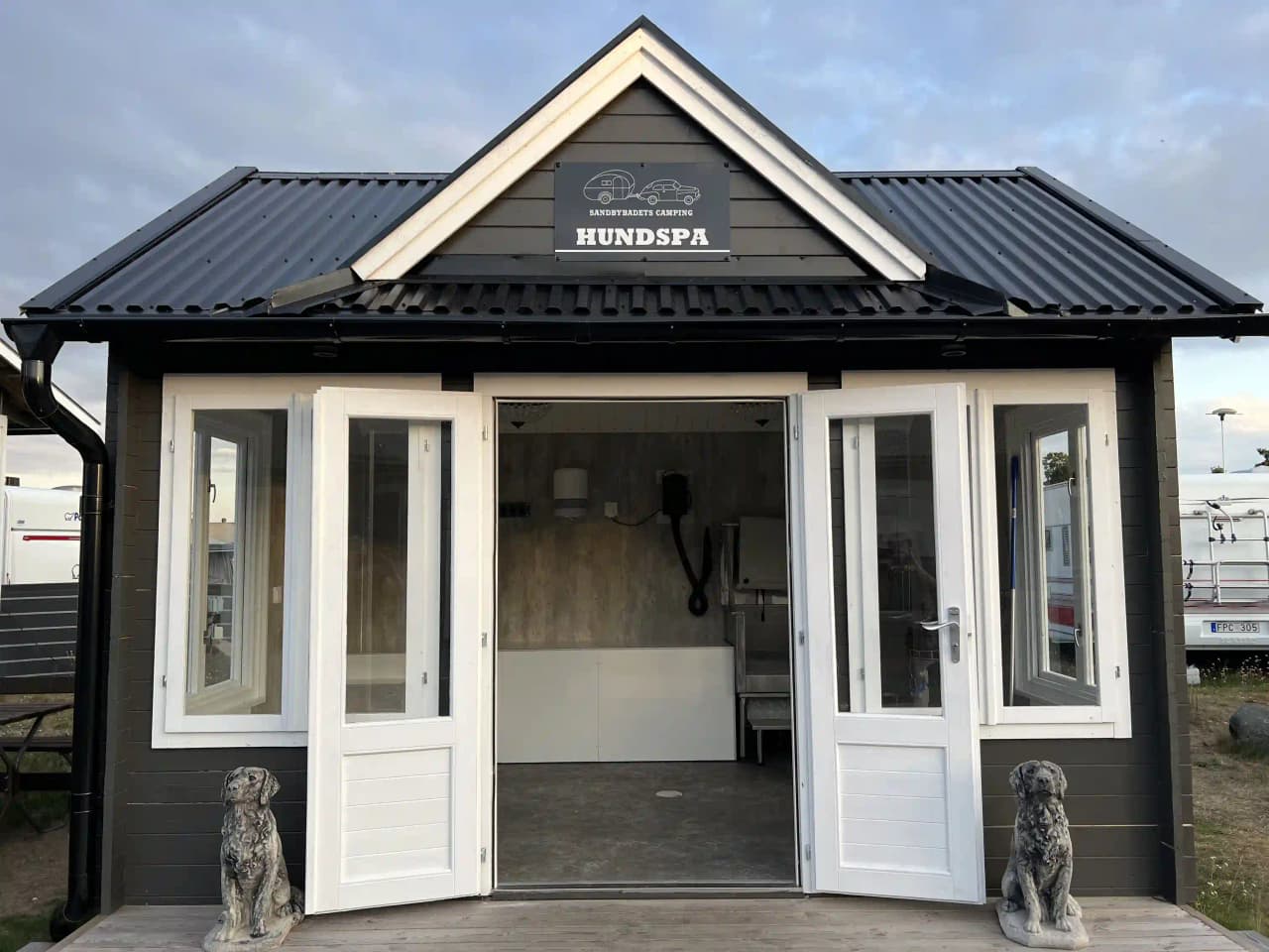 En enkel träbyggnad vid Sandbybadets camping, med ett tak av metall och en vit dörr. Den står i en naturskön miljö och används möjligen som ett förråd eller kiosk.