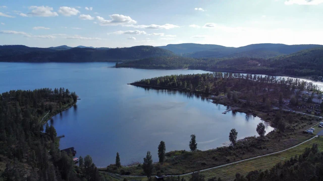En naturskön vy över Måvikens camping & naturområde, med ett blått vattendrag omgivet av grönskande natur och höjder. Moln svävar ovanför, vilket ger en rofylld atmosfär.