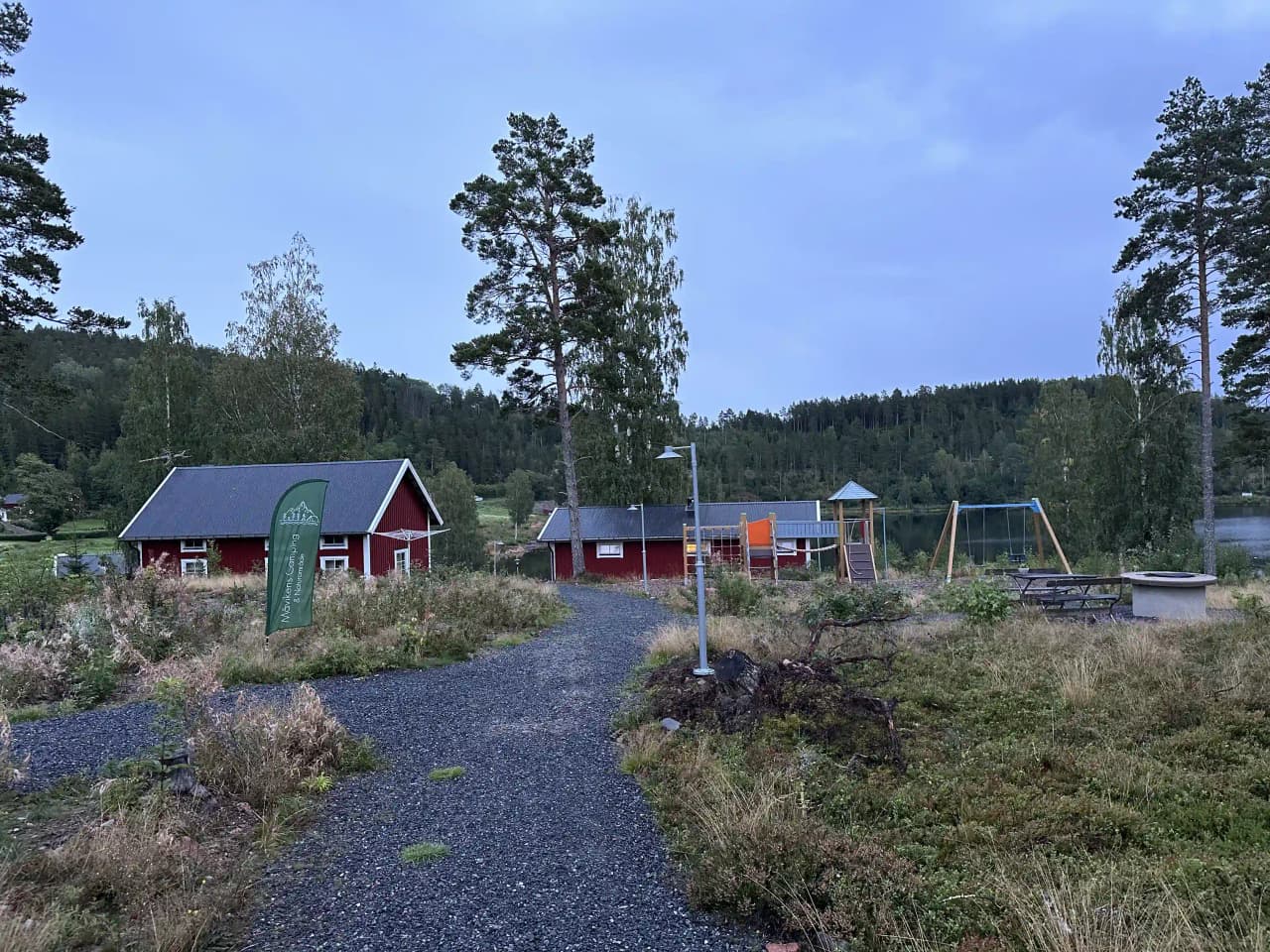 En naturskön campingplats omgiven av träd och grönska, med en stuga som syns i bakgrunden och en gräsbevuxen äng i förgrunden.