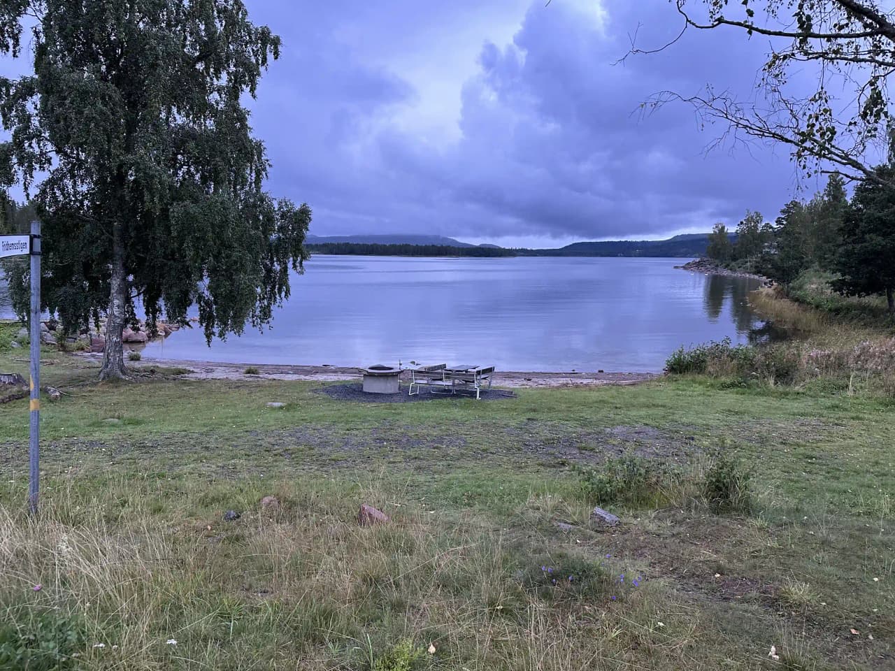 En vy över Måvikens camping och naturområde med ett stort blått vattenområde omgärdat av grönskande kustlinje. Solen lyser på det glittrande vattnet under en himmel med vita moln.