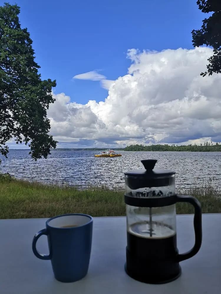 En kopp på ett bord med en sjö och molnig himmel i bakgrunden, solen lyser över vattenytan och stranden vid Hägern Bed & Nature.