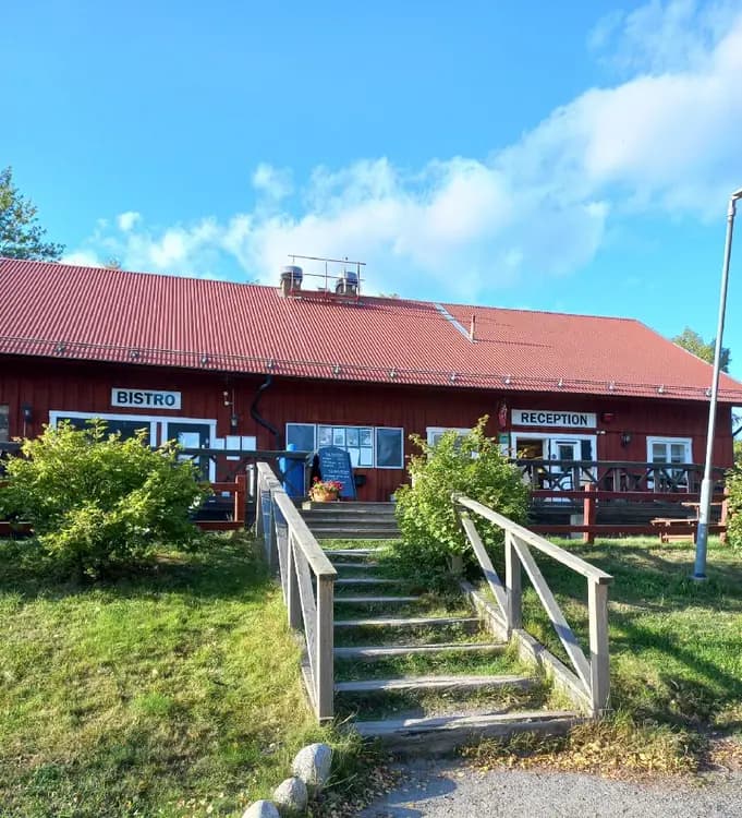 En träbänk utomhus vid ett gålö havsbad. Bänken står nära ett serveringsområde som liknar en restaurang eller värdshus. Det finns utomhusmöbler och platsen verkar vara en viloplats för besökare. I bakgrunden syns en byggnad med träpanel.