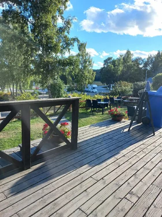 En solig dag vid Gålö Havsbad med trädeck som sträcker sig mot vattnet. Utomhusmöbler i trä står på däcket, omgivna av grönska och skugga. En gångväg av plankor leder besökare längs stranden.