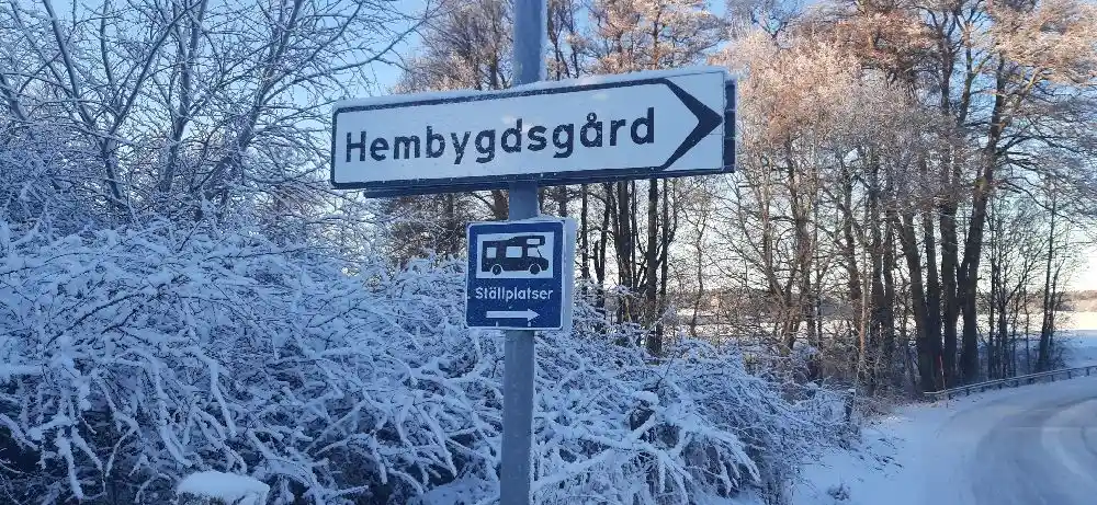 En snötäckt väg leder till Kvarngården Hembygdsgård, omgiven av frostiga grenar och träd. En skylt med platsens namn står vid sidan av vägen.