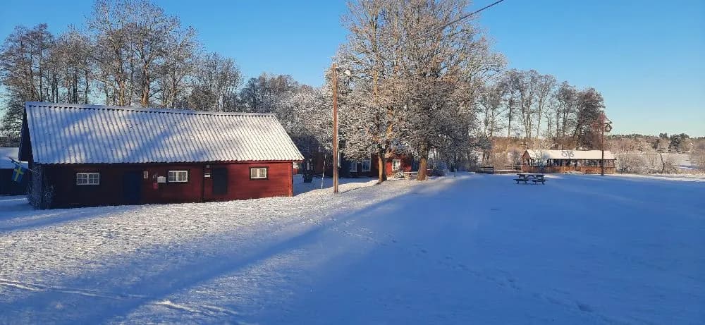En vinterklädd kvarngård omgiven av snötäckta träd och grenar, med ett traditionellt trädfodrat hus och snö på taket.