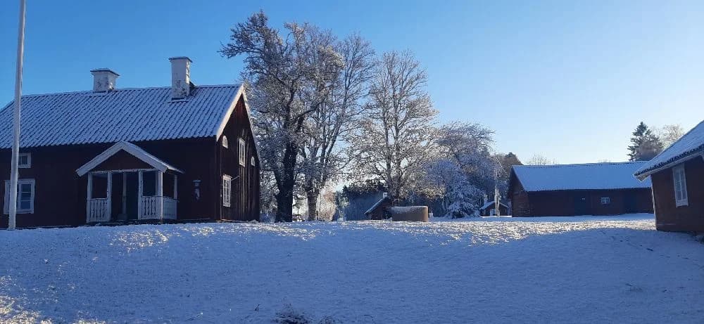 En vintervy av Kvarngården hembygdsgård, där en pittoresk byggnad täcks av snö. Nakna grenar sträcker sig ut mot den frusna himlen, vilket skapar en charmig och nostalgisk stämning på den historiska platsen.