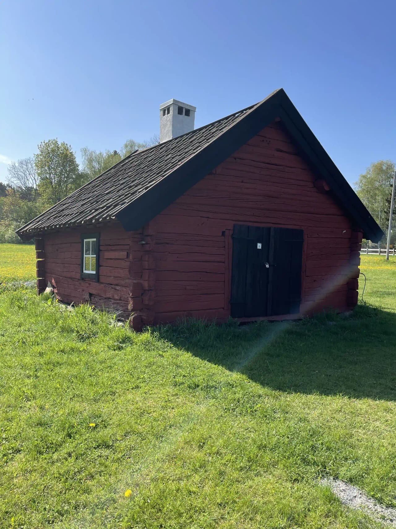 En rustik träbyggnad med grästak, omgiven av frodig gräsyta. Bakom byggnaden skymtar ytterligare mindre hus i samma traditionella stil. Landskapet är öppet och lantligt, vilket understryker den historiska charmen hos Kvarngården Hembygdsgård.
