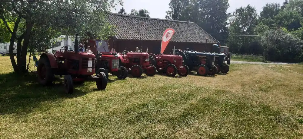 En gammal traktor står parkerad vid Kvarngården Hembygdsgård, omgiven av grönska. Traktorn har stora hjul med mönstrade däck och är ett typiskt exempel på äldre jordbruksmaskineri.
