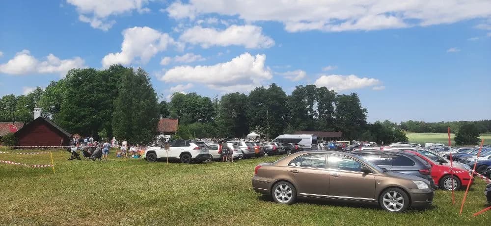 Bild av kvarngården hembygdsgård med bilar parkerade i förgrunden på en grusplan. Byggnaden och en lantlig miljö syns i bakgrunden.