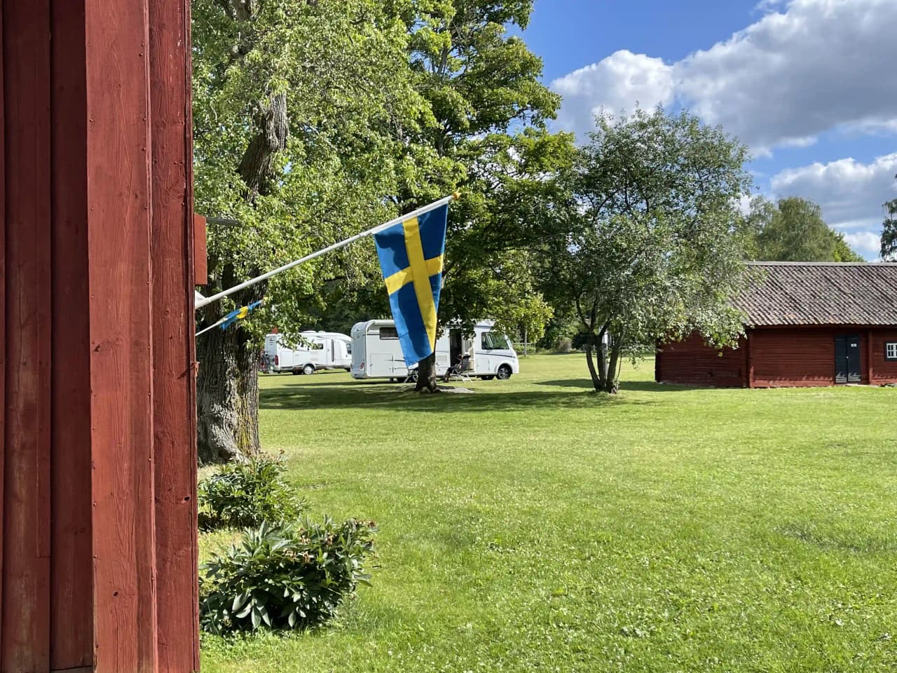 En grönskande hembygdsgård med gräs och ängsblommor, där en liten stuga står vid sidan av en pol och en öppen gräsmatta.