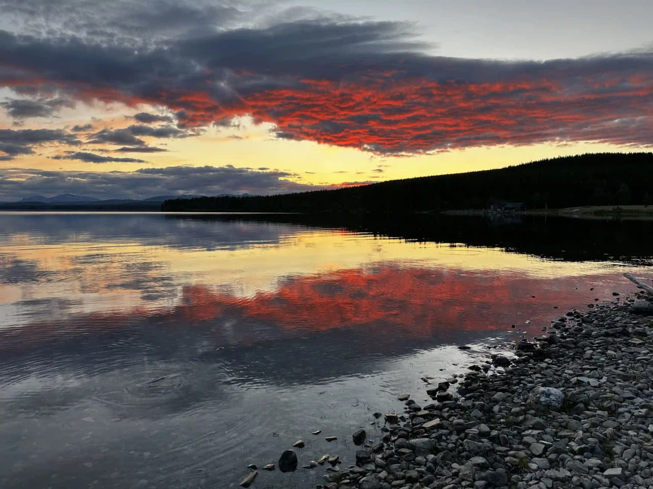 En rofylld sjö med blått vatten sträcker sig ut mot horisonten under en vackert färgad kvällshimmel, där mjuka moln belyses av den nedgående solens glöd.