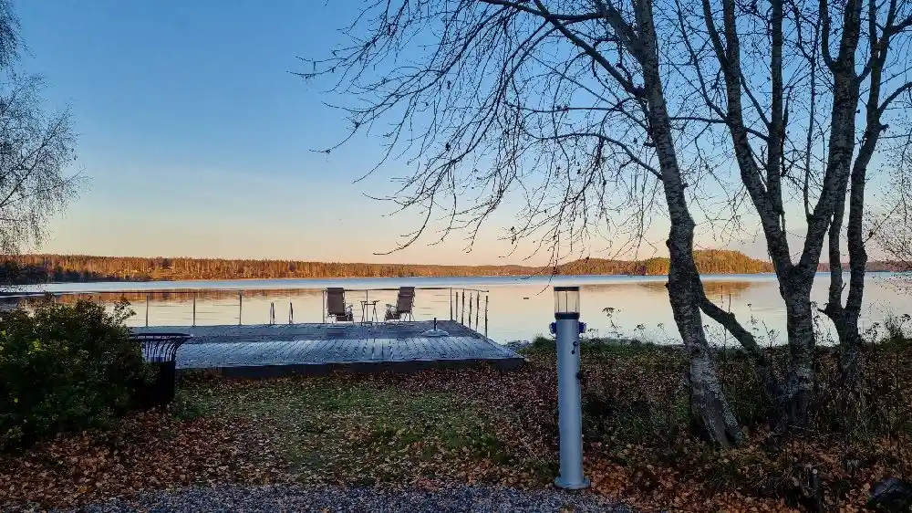 En liten sjö med klart blått vatten omgiven av skog och grönska under en klarblå himmel. Solen lyser över landskapet och skapar en fridfull atmosfär vid strandkanten.