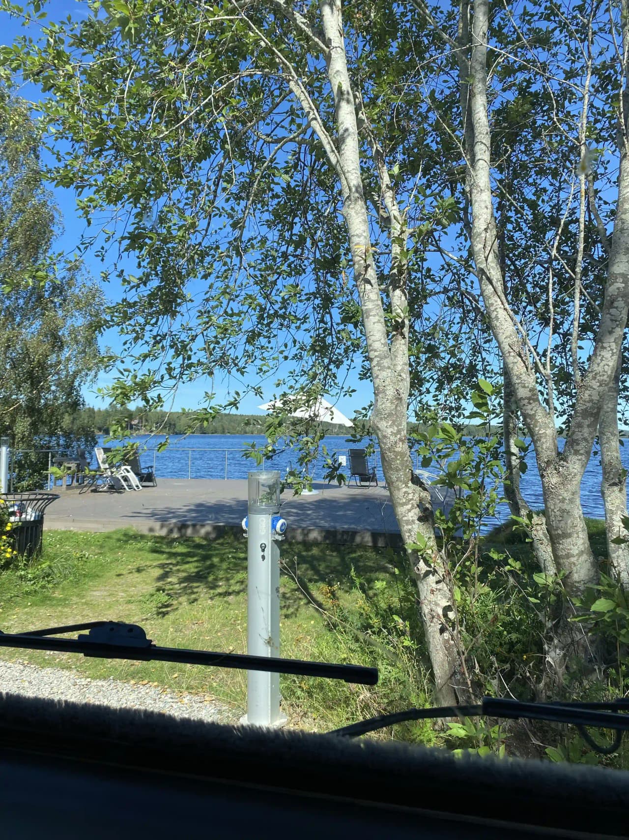 En bil står parkerad under en björk vid Sagaliden, omgiven av skuggande trädgrenar.