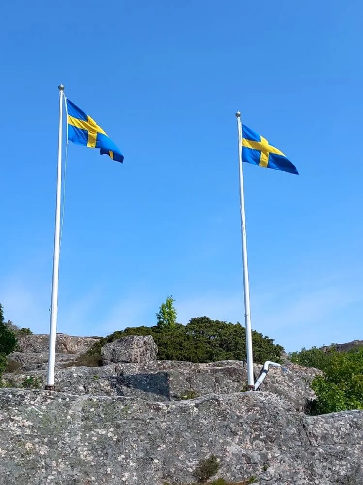 En blå flagga vajar i vinden mot en klar himmel vid Fjällbacka camping, med solljus över klipporna i bakgrunden.