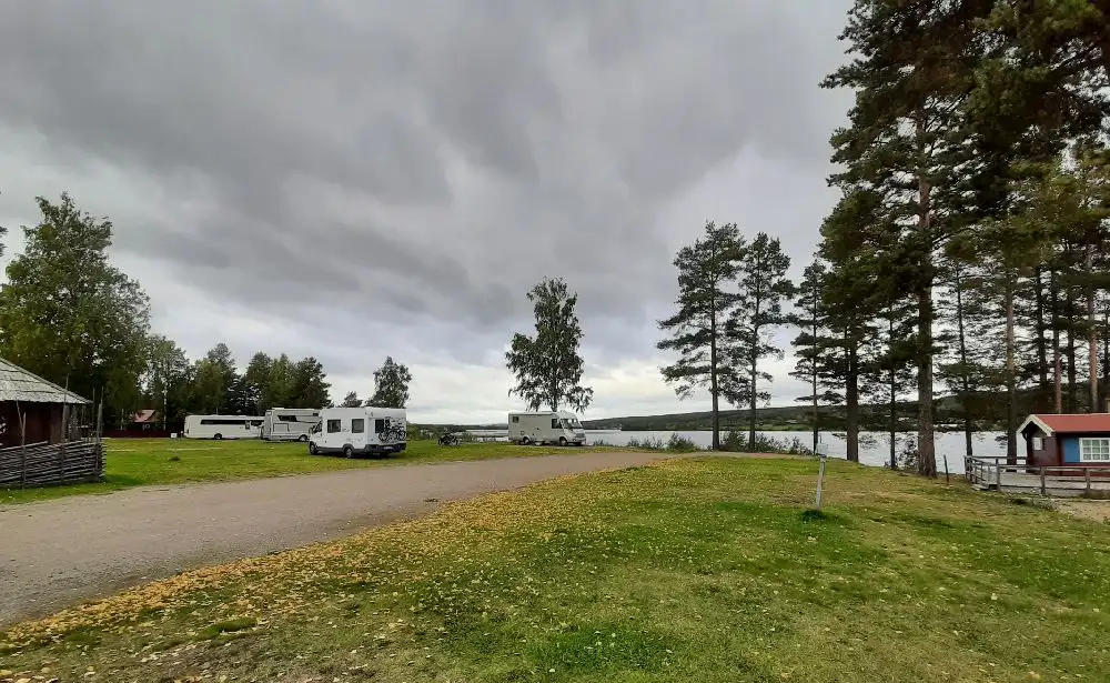 En solig dag vid Ljusdals camping med husbilar parkerade på en grön gräsyta, omgiven av lummiga träd och under en himmel täckt av fluffiga cumulusmoln.
