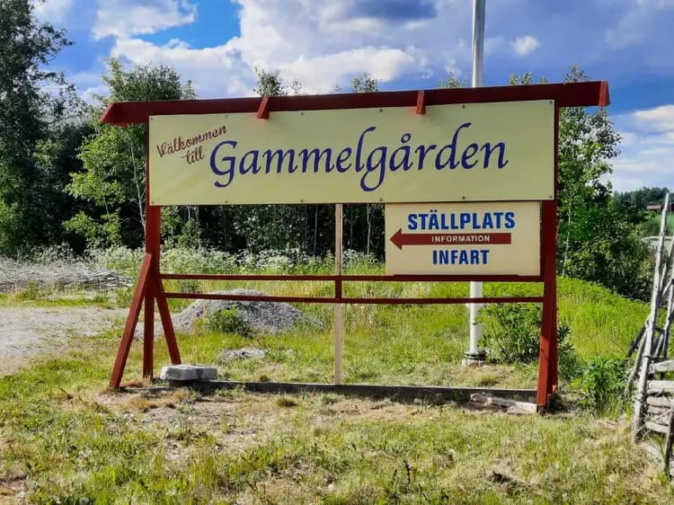 Bild på Västerfärnebo hembygdsgård: En grusad stig omgiven av gräs leder fram till en skylt med namnet på platsen. Skogsområde syns i bakgrunden under en klar himmel.