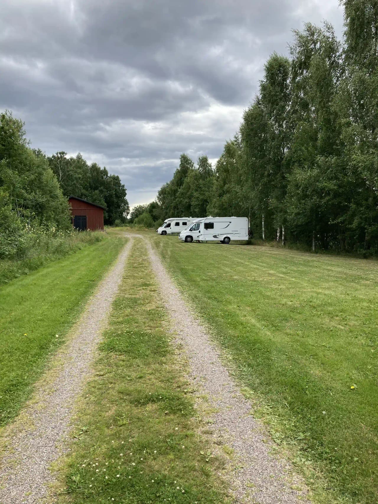 En grönskande hembygdsgård i Västerfärnebo med träd och gräsmarker. En stig slingrar sig genom landskapet där en husvagn syns i bakgrunden, placerad nära en gårdsplan.