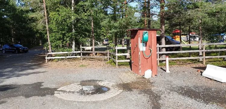 En bild från Habo Camping och Stugby visar en välordnad gårdsplan med parkerade fordon, inklusive en SUV och en småbil, omgivna av grönskande trädgård och små trädgårdsbyggnader som skjul. En grusad stig leder genom området.