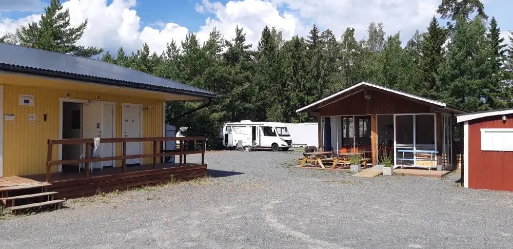 Bild av Habo Camping och Stugby med en stuga omgiven av trädgårdsbyggnader. Framför stugan syns en parkering med en SUV och bakljuset på bilen syns tydligt.