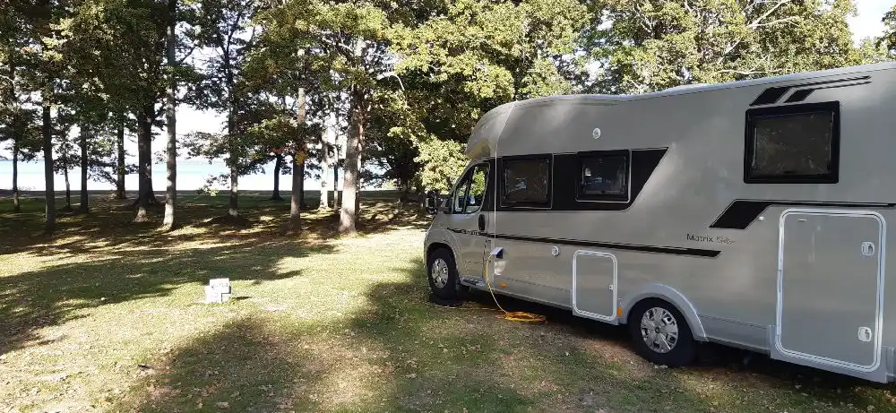 En husvagn och bil parkerade på Hjälmargården & Läppe Camping, omgiven av grönska under en klar himmel.