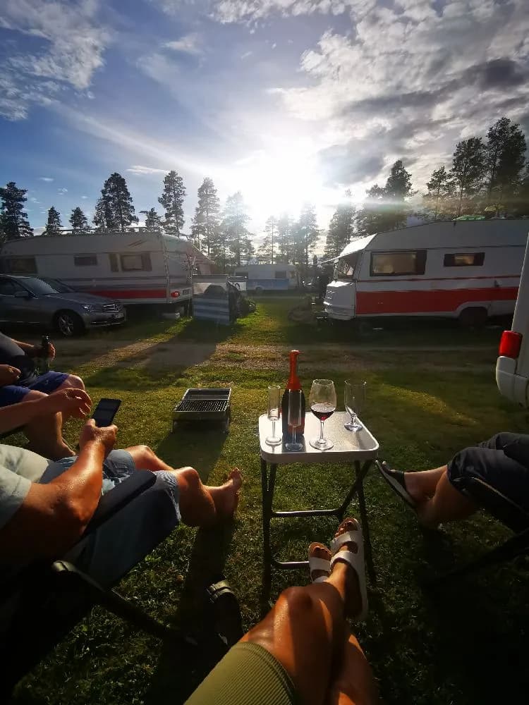 En solig dag vid Bovikens havsbad med husvagnar och husbilar parkerade på platsen. Campingstolar och fritidsfordon står uppställda på gräset medan solens strålar skapar en lätt linsöverstrålning.