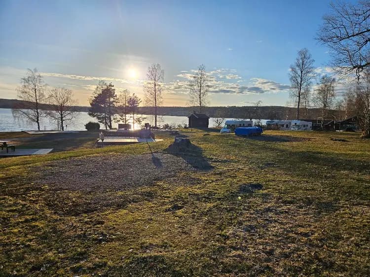 En fridfull campingplats vid en sjö, med gröna gräsmattor och skuggor som sträcker sig över marken i det mjuka morgon- eller kvällsljuset.