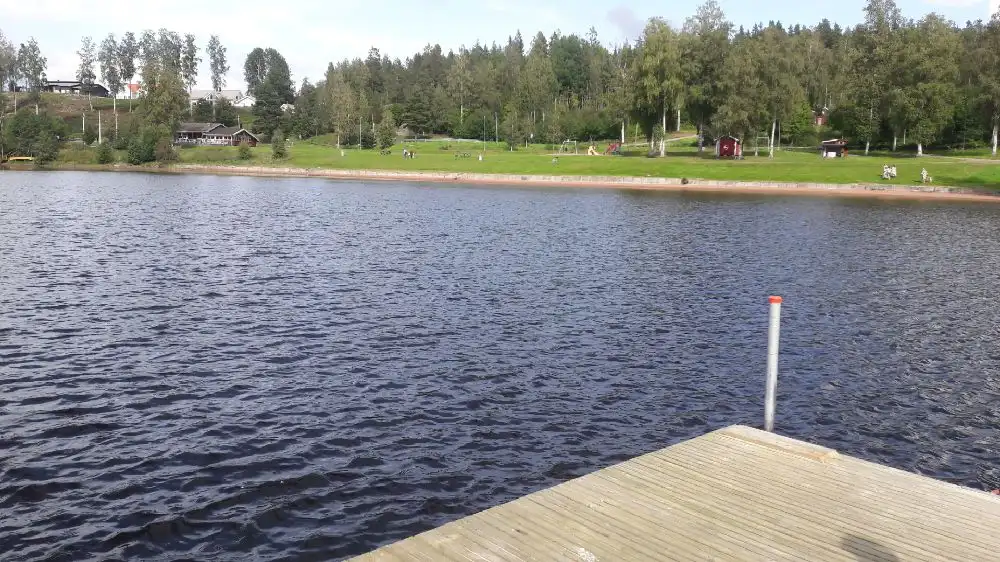 En vy över Säters camping med en sjö i förgrunden och skogsklädda kullar i bakgrunden. Vattenytan är lugn och omgivningen erbjuder möjligheter för rekreation och naturupplevelser.