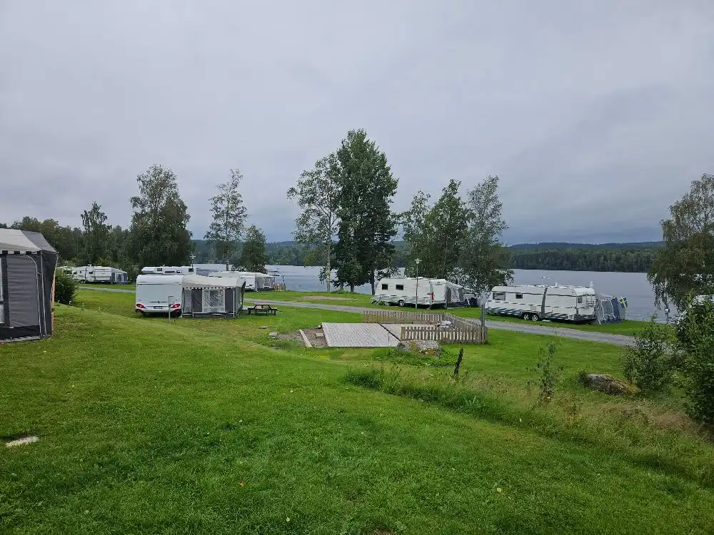 En naturskön vy över Säters camping med en sjö omgiven av grönskande landskap och moln på himlen. Perfekt för avkoppling och friluftsliv.