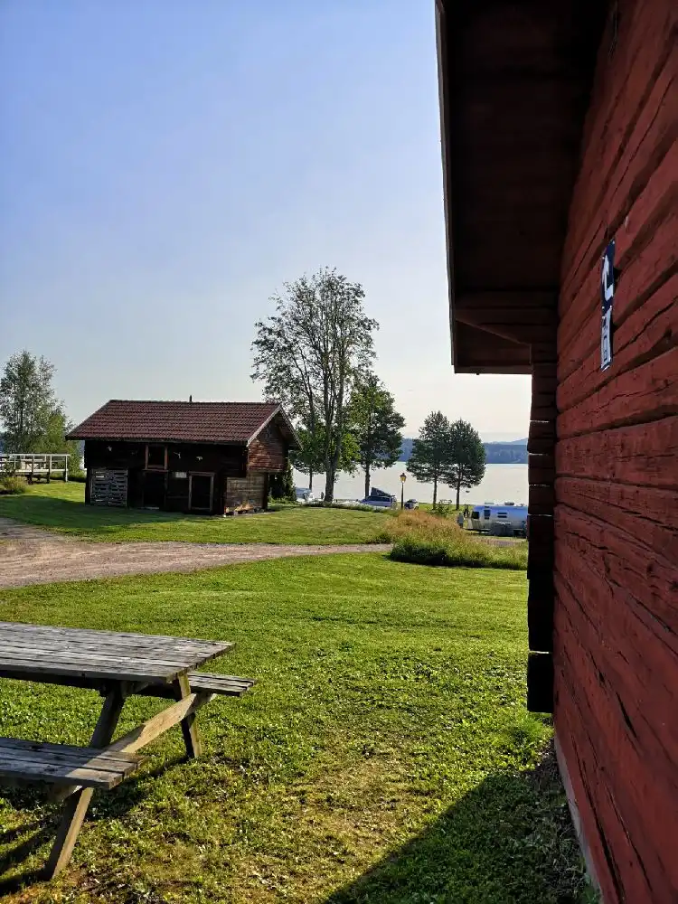 Bild på Västanviksbadets Camping i Leksand, med en grön gräsmatta i förgrunden, omgiven av flera små stugor i lantlig miljö. Tak och lador syns i bakgrunden, vilket skapar en hemtrevlig atmosfär.