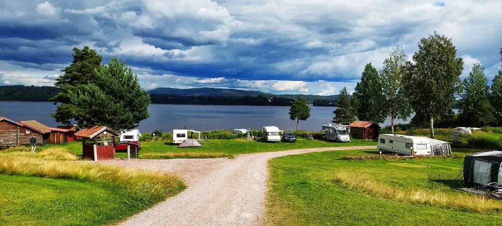 En naturskön campingplats vid Västanviksbadet i Leksand, omgiven av kullar och molntäckt himmel, med vacker utsikt över en sjö och dess kustlinje.