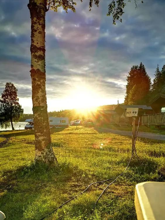 Bild av Snibbens camping med soluppgång över ett somrigt landskap. Solens strålar lyser upp den gröna gräsmattan och den omkringliggande naturen i ett lantligt område.