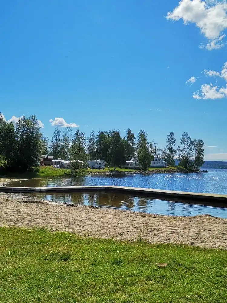 Bild av Snibbens camping: En somrig vy med gröna träd, en gräsmatta och skuggade områden vid kanten av en sjö eller damm. Perfekt för avkoppling och friluftsliv.