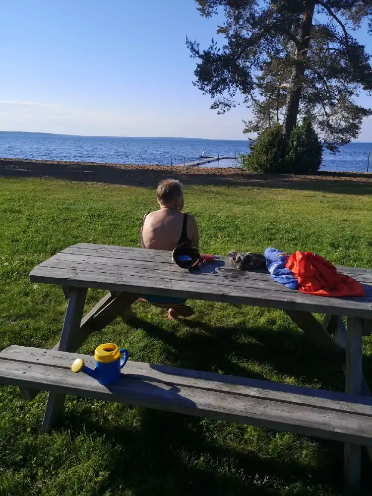 En träbänk och ett picknickbord står på en gräsmatta vid Otterbergets bad och camping. I bakgrunden syns havet under en klar himmel, vilket ger en avkopplande och naturskön atmosfär perfekt för semester och rekreation.