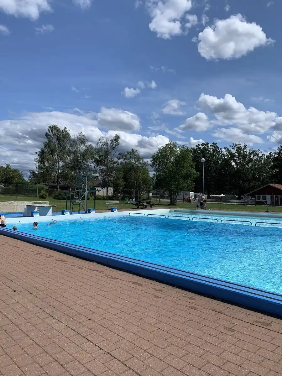 Bild med en utomhuspool vid Kosta Bad och Camping, omgiven av en grönskande parkmiljö. Flera människor syns njuta av semestern i solen, några simmar i poolen medan andra kopplar av under skuggan av parasoller.