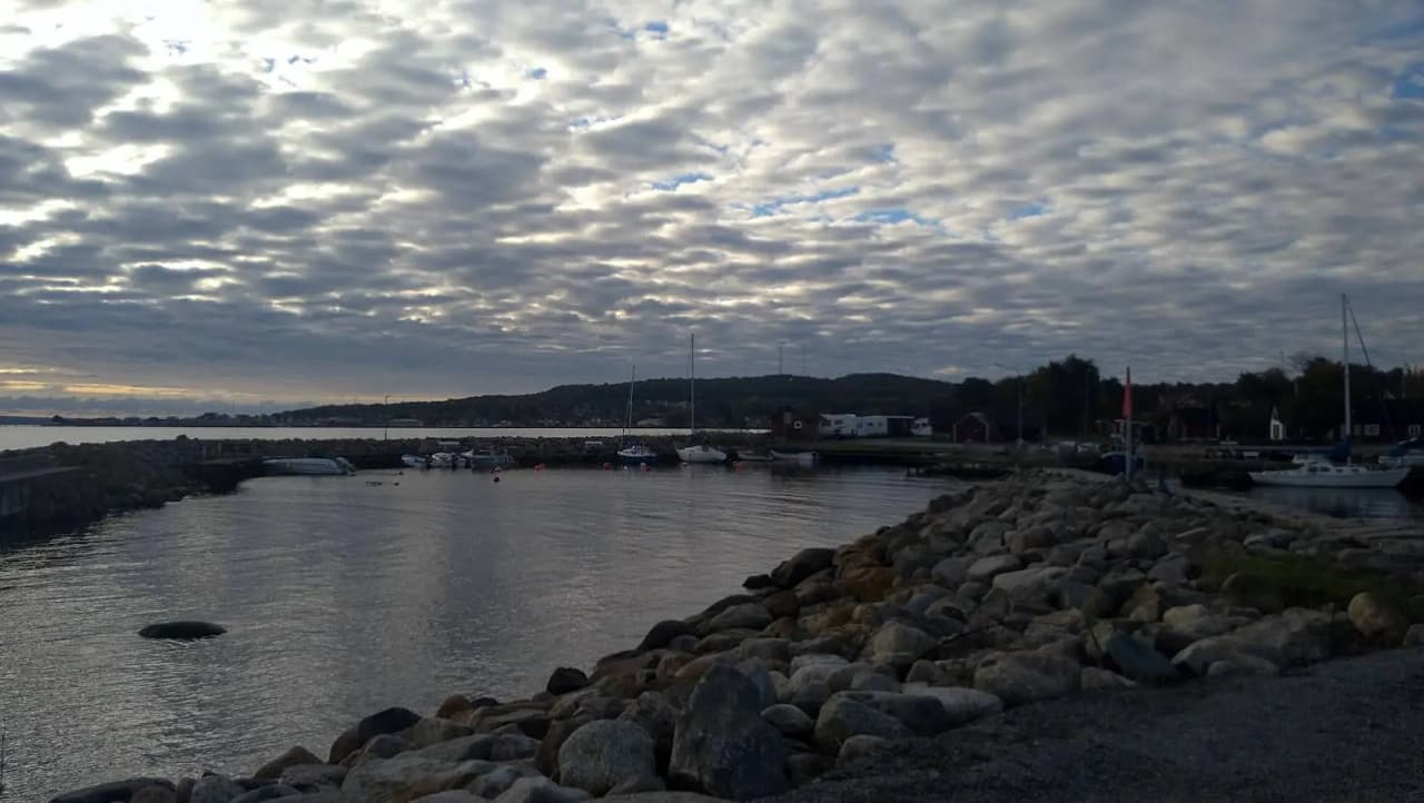 En naturskön vy vid Valjevikens camping med en sjö i förgrunden, omgiven av kustlinje med klippor och grönska under en molnig himmel.