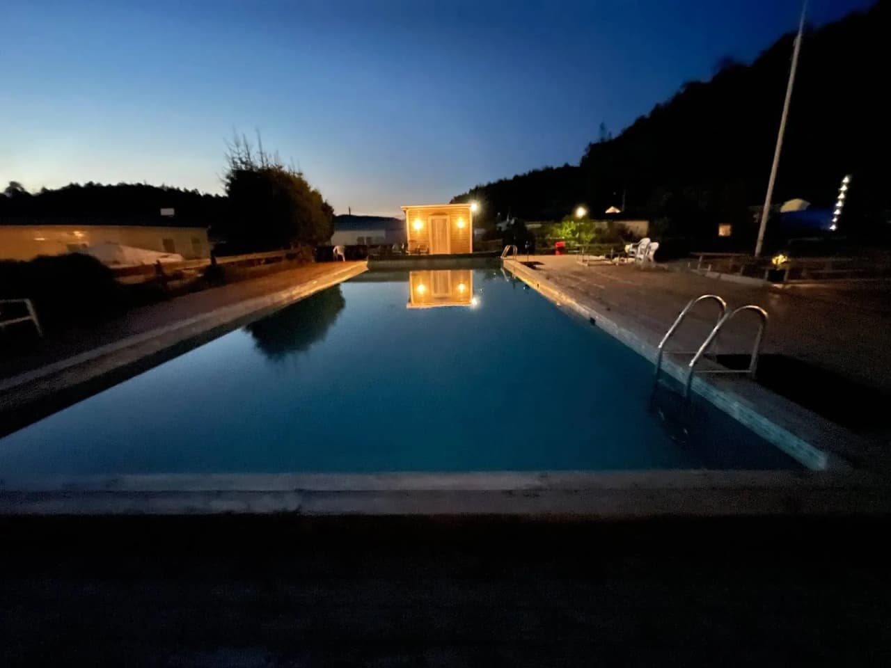 En nattbild av en swimmingpool på Ylserød Camping, omgiven av lyktor och speglingar i vattnet, vilket skapar en lugn och avkopplande atmosfär.