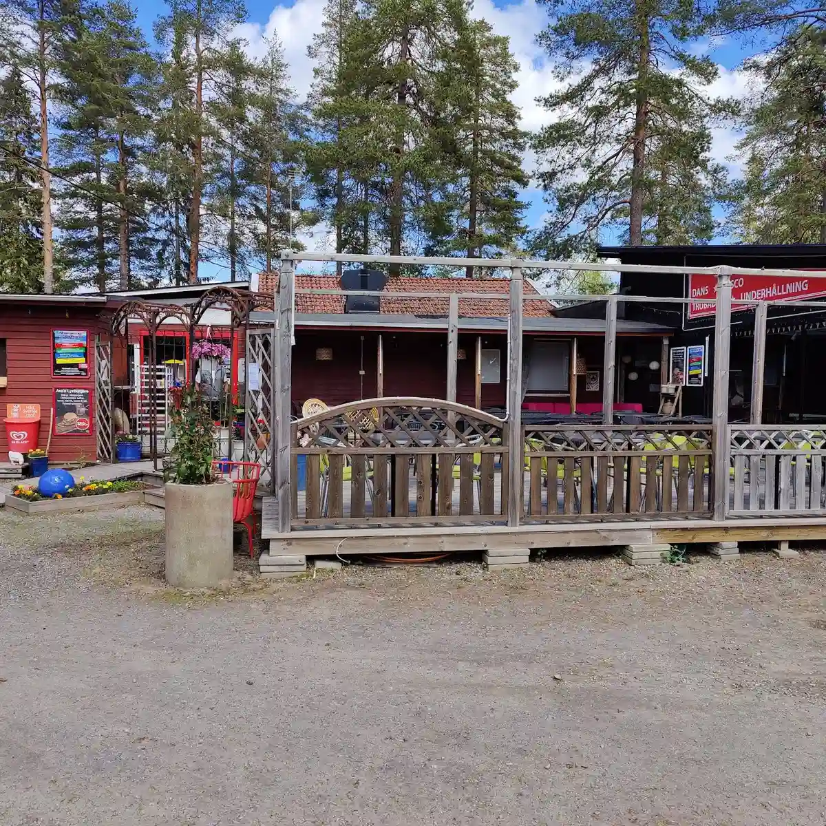 En solig dag vid Ava havsbad och camping. Ett öppet område med parkering och flera utemöbler såsom bord och bänkar i skuggan. Flera utomhusstrukturer och en veranda syns i bakgrunden, vilket skapar en inbjudande vila och samlingspunkt.