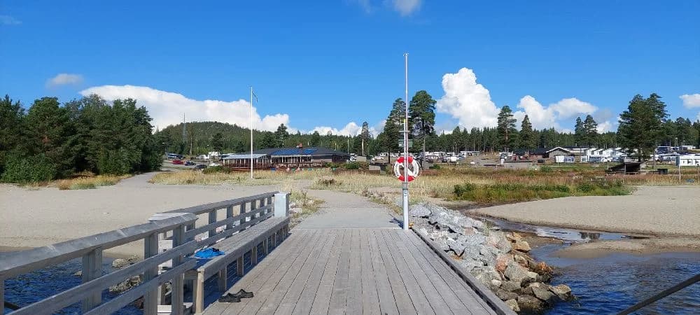 En solig dag vid Gullviks Havsbad Camping & Stugby med en träpromenad omgiven av tallar och andra barrträd. I förgrunden syns ett staket och en skuggig gångväg.