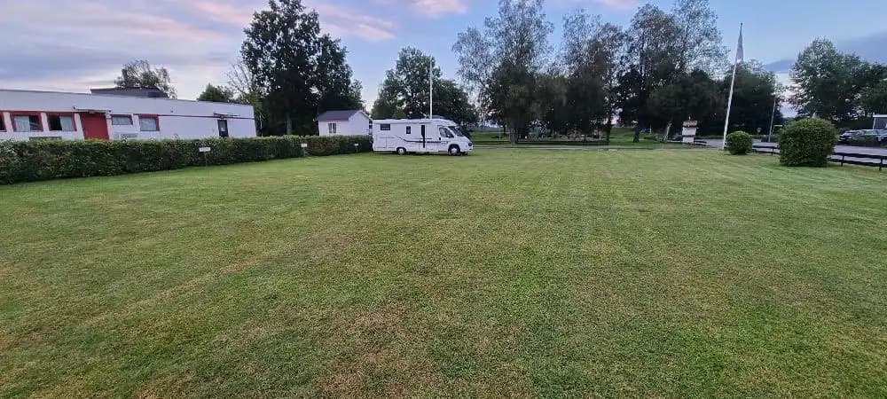 En grön campingplats med gräsmatta och flera husvagnar uppställda. Flera campare njuter av omgivningen i sjöparkens camping.